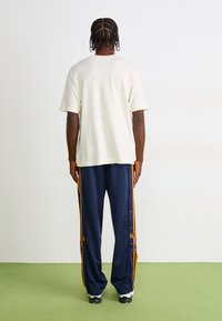 En person med fletninger står overfor en hvid væg iført en simpel off-white T-shirt, navy joggebukser med orange striber og hvide sneakers.