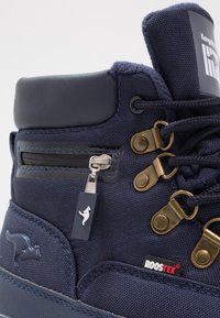 Bottine en tissu bleu marine avec accents en cuir, œillets métalliques et fermeture éclair décorative. Comprend un logo et des surfaces texturées.