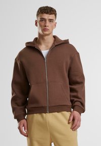 Brauner Reißverschluss-Hoodie aus weichem Stoff, mit einer großen Kapuze, Fronttaschen und gerippten Bündchen. Kombiniert mit hellgelben Jogginghosen.