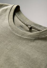 Olive roheline puuvillase t-särgi kaeluse lähivõte, kus on nähtav bränditekst ja kangatõmblus neutraalsel taustal.