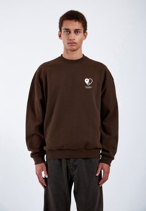 Bruine oversized trui met een ronde halslijn, geribbelde mouwen en zoom, met een witte hartgrafiek en tekst op de linkerborst.