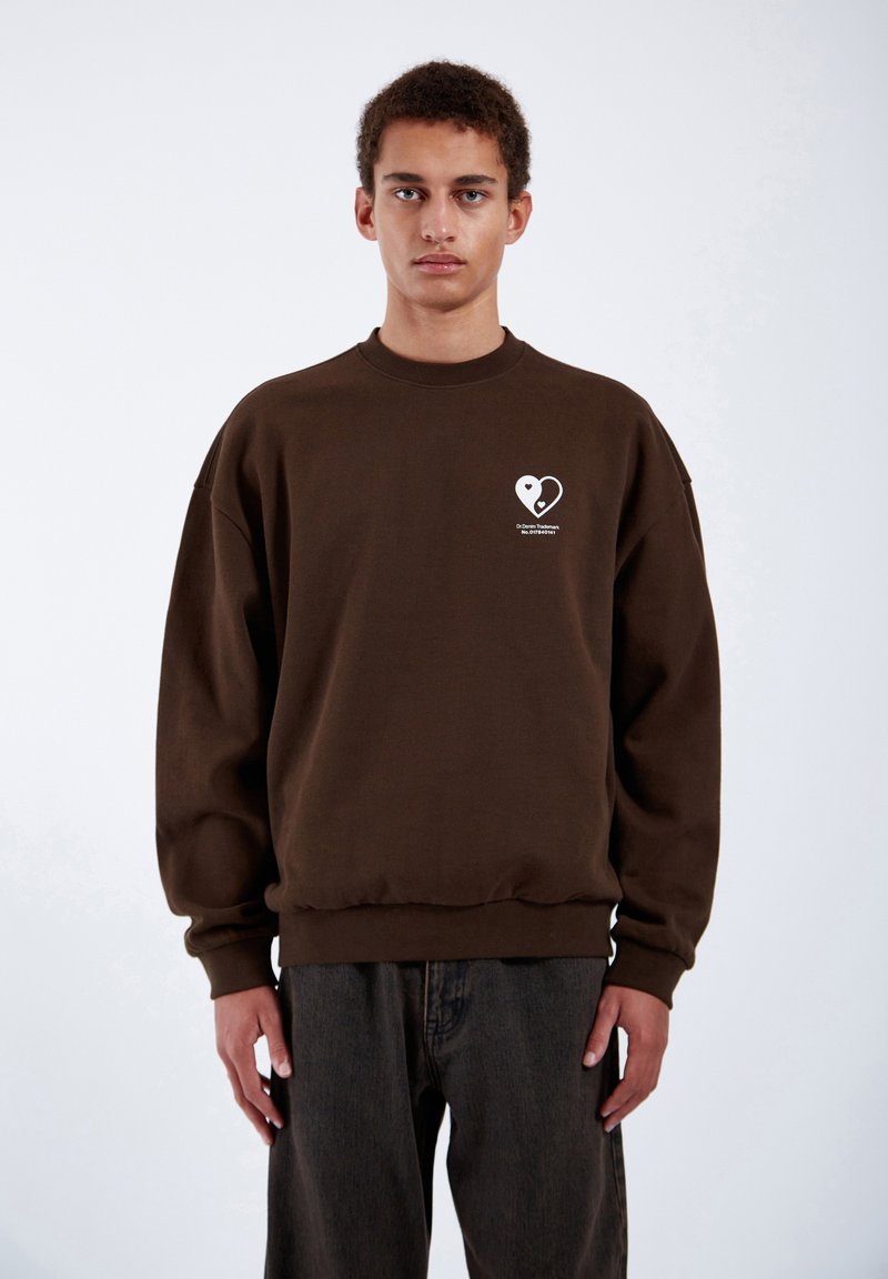 Sweatshirt marrone oversize con scollo rotondo, polsini e orlo a costine, caratterizzato da una grafica a cuore bianco e testo sul petto sinistro.