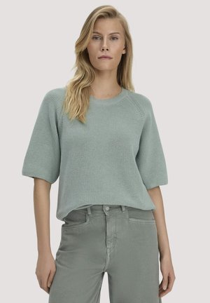 Vrouw met lang blond haar, gekleed in een lichtgroene gebreide trui met ellebooglange mouwen en bijpassende hoge taille broek.