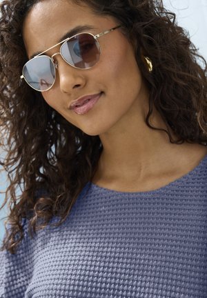 Gold-framed sunglasses with brown lenses, worn by a person with curly hair. The textured, purple top features a round neckline.