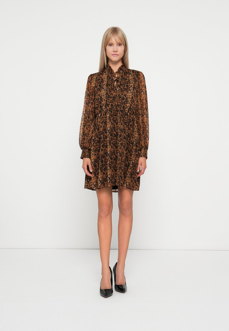 Brun leopardmønstret kjole med lange ærmer, knaplukning foran og lagdelt design. Brukt med svarte høye hæler, på en ensfarget bakgrunn.