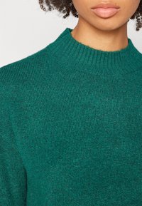 Groene gebreide trui met een hoge, geribbelde kraag en een textuurstof. Het ontwerp toont een close-up van het lichaam.