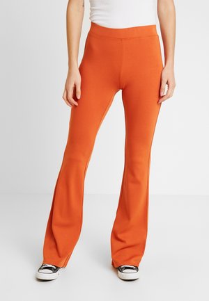 Pantalons évasés orange vif, extensibles, avec une taille haute et une texture lisse, associés à des baskets noir et blanc.