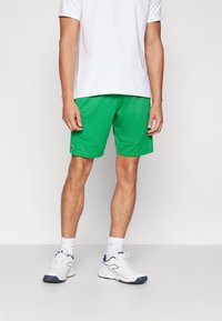Shorts sportivi verdi realizzati in tessuto traspirante, con orlo testurizzato, tasche laterali e logo. Abbinati a una maglietta bianca e scarpe da ginnastica.