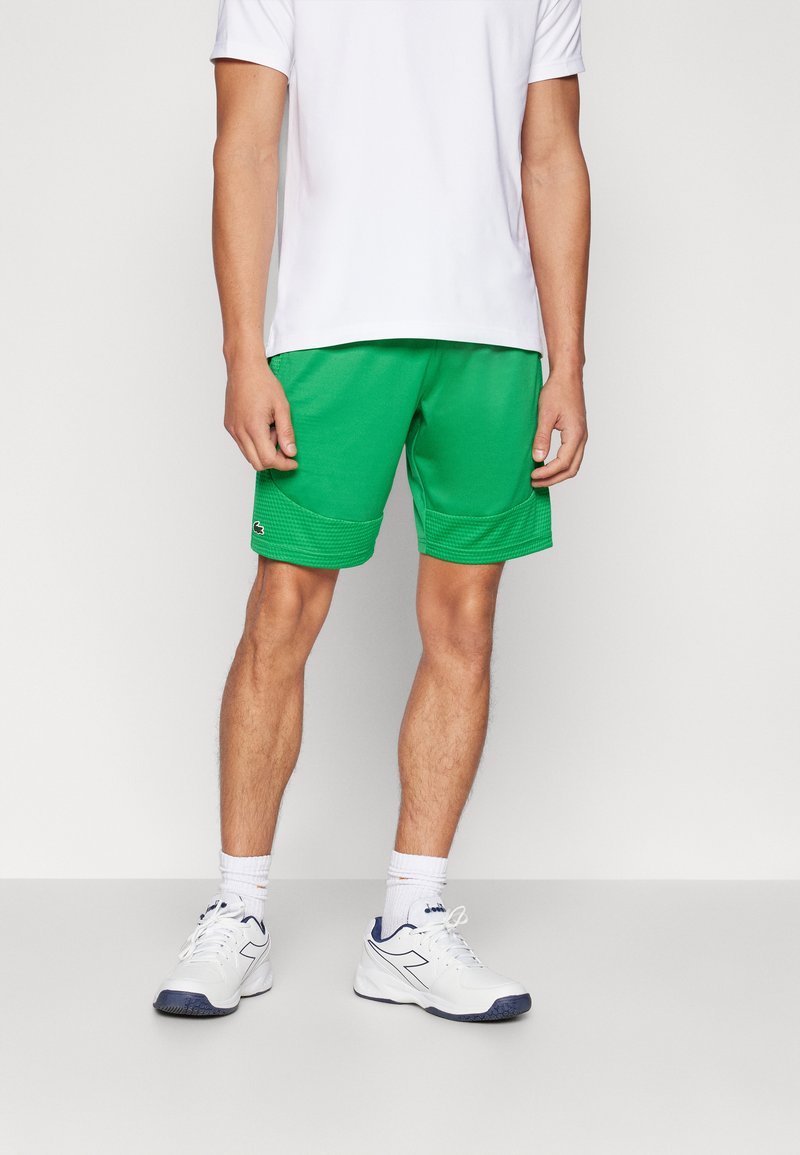Shorts sportivi verdi realizzati in tessuto traspirante, con orlo testurizzato, tasche laterali e logo. Abbinati a una maglietta bianca e scarpe da ginnastica.