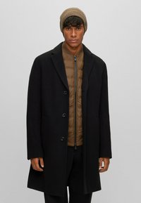 Cappotto lungo nero in lana con colletto a punte e cinque bottoni sopra una giacca imbottita marrone a trapuntatura; berretto beige a costine.