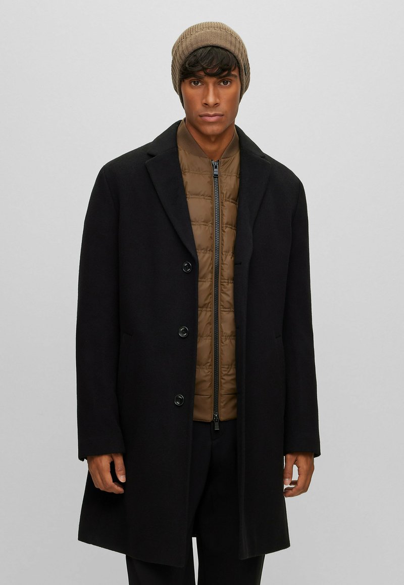 Cappotto lungo nero in lana con colletto a punte e cinque bottoni sopra una giacca imbottita marrone a trapuntatura; berretto beige a costine.