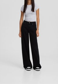 Jeans negros de pernera ancha con una cintura alta y diseño de cinco bolsillos. Combinados con una camiseta gris ajustada de manga corta y zapatos en blanco y negro.