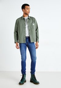 Zielona koszula z guzikami noszona na szarym t-shircie, zestawiona z niebieskimi dżinsami slim-fit i zielonymi butami na sznurówki, stojąc na neutralnym tle.