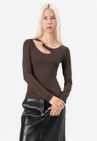 Top marrone a maniche lunghe con un taglio vicino alla spalla e un accento metallico decorativo. Tessuto liscio, design aderente. Abbinato a una gonna in pelle nera.