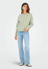 Vrouw in een lichtgroen sweatshirt met de tekst "ENJOY", lichtblauwe wijde jeans en witte puntige schoenen, staande tegen een effen achtergrond.