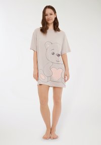 Giovane donna in piedi a piedi nudi, che indossa un vestito t-shirt beige oversize con una grande grafica di orso di peluche che tiene un cuore rosa.