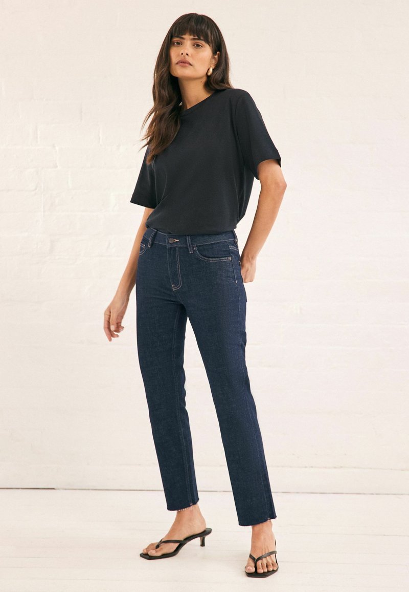 Top negro de manga corta combinado con jeans ajustados de color azul oscuro que cuentan con bolsillos frontales y costuras en contraste, complementado con sandalias de tacón negras.