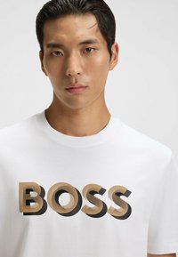 Młody mężczyzna z krótkimi ciemnymi włosami, ubrany w białą koszulkę z napisem "BOSS" w dużych beżowych i czarnych literach.