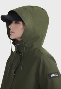 Veste imperméable verte avec capuche, cordons de serrage réglables et fermeture éclair sur le côté. Dotée d'un écusson noir sur la manche gauche.