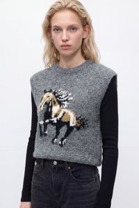 Jeune femme aux cheveux blonds ondulés porte un pull sans manches gris avec un motif de cheval multicolore sur une chemise noire à manches longues et un jean noir.