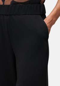 Un pantalon noir présente un tissu lisse et texturé avec une ceinture extensible et des poches latérales, montrant un gros plan de la taille et de la poche.