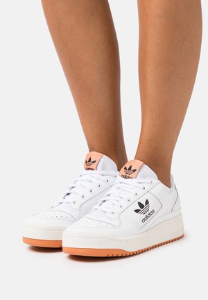 Weiße Adidas-Sneaker mit pfirsichfarbenen Akzenten, getragen an nackten Beinen, mit schwarzem Adidas-Logo und dickerer pfirsichfarbener Sohle auf hellem Hintergrund.