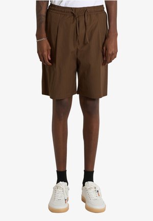 Persona che indossa pantaloncini marroni a righe fino al ginocchio, calzini neri, scarpe da ginnastica bianche e una camicia bianca, in piedi davanti a uno sfondo uniforme.