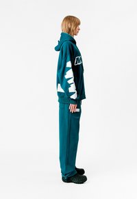 Sudadera de gran tamaño en color verde azulado con estampado gráfico en blanco en las mangas, combinada con pantalones de chándal a juego y calzado negro texturizado. Corte simple y relajado.