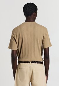 Uomo con capelli ricci corti che indossa una maglietta a maniche corte beige a coste infilata in pantaloni khaki chiari con una cintura intrecciata scura, di spalle.