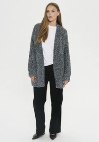 Žena s dlhými hnedými vlasmi oblečená v šedej hrubej pletenej kardigane, bielej blúzke, čiernych širokých nohaviciach a čiernych špicatých topánkach, stojí.