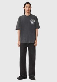 Melna oversized t-kreklā ar grafisku zīmējumu uz krūšu, kombinācijā ar melniem platās kājas džinsiem un melnā kedām, uz neitrāla fona.