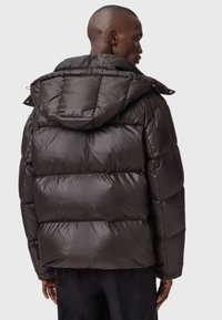 Mand iført en mørkebrun puffjakke med en stor hætte, set fra ryggen mod en ensfarvet lys baggrund.