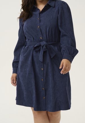 Woman wearing a navy blue corduroy button-up dress with long sleeves and a tied waist belt, standing against a plain background.