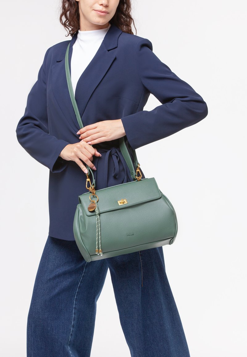 Grøn læderhåndtaske med struktureret overflade, gyldent hardware og en aftagelig rem, holdt af en person i en navy blazer og hvid top.