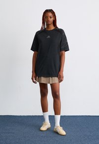 Mlada ženska s pletenicami, oblečena v oversized črno majico Adidas, bež plisirano krilo, bele nogavice in bež teniske, stoji na modrem preprogi.