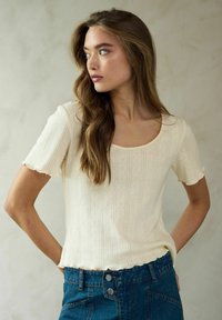 Krämfärgad kortärmad topp med rundad halsringning, strukturerade vertikala ränder och scallopade ärmslut, tillsammans med höga jeans.