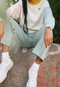Sudadera blanca con mangas en tonos pastel rosa y azul, cuello amarillo. Pantalones de pierna ancha en verde claro. Zapatillas blancas con logo y suela texturizada.