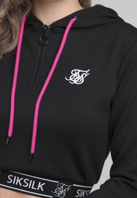 Haut à capuche noir court avec une fermeture éclair et des cordons de serrage roses. Équipé d'un logo blanc à l'avant et d'une ceinture noire et blanche avec logo.