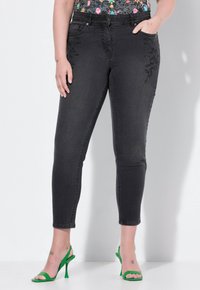 Jeans de denim negro con un diseño ajustado, que presentan bordados florales en los bolsillos delanteros y una textura suave. Llevados con sandalias de tacón verdes.