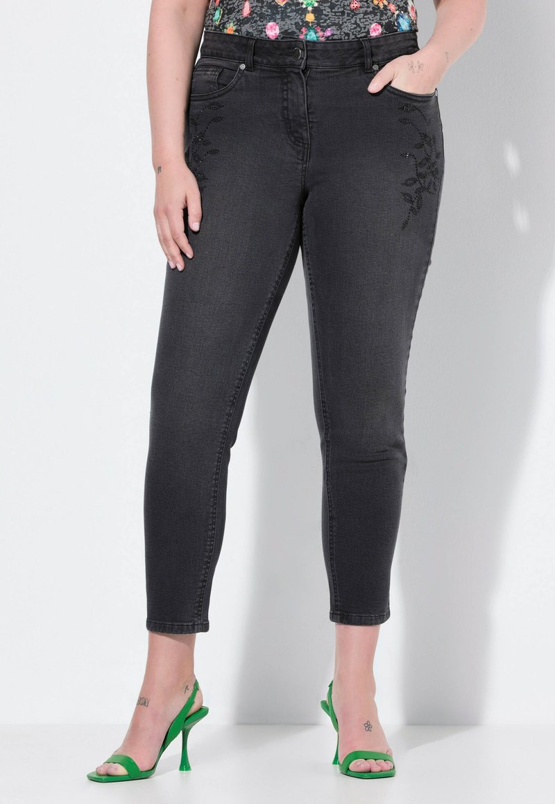 Jeans de denim negro con un diseño ajustado, que presentan bordados florales en los bolsillos delanteros y una textura suave. Llevados con sandalias de tacón verdes.