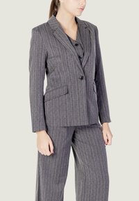 Completo grigio a righe con giacca su misura e pantaloni a gamba larga. Presenta un collo a pattina, due tasche frontali e dettagli di chiusura con bottoni.