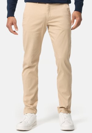 Homme portant un pantalon beige slim-fit, des baskets blanches et une chemise bleu marine à manches longues, debout devant un fond blanc uni.