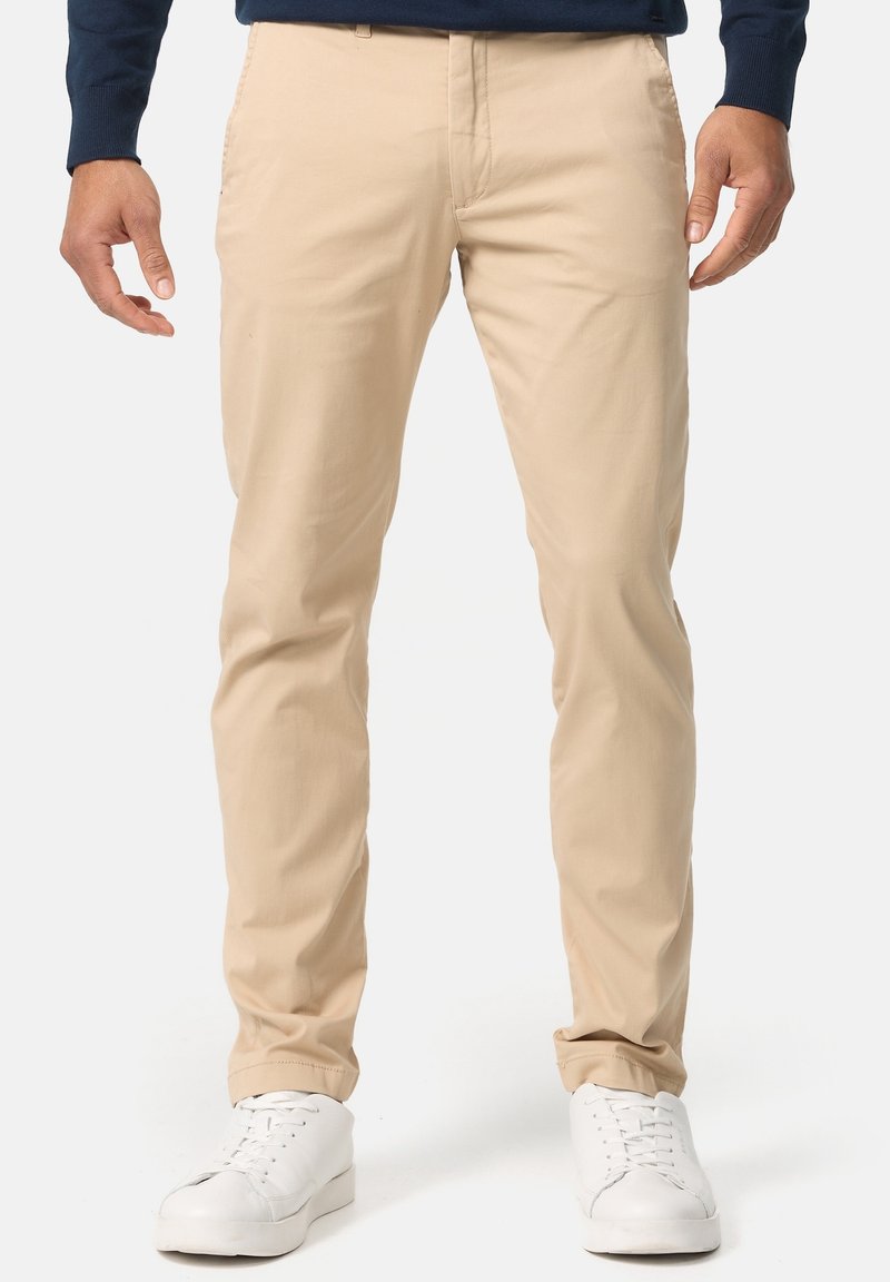 Homme portant un pantalon beige slim-fit, des baskets blanches et une chemise bleu marine à manches longues, debout devant un fond blanc uni.