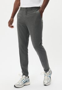 Muž v šedých slim-fit kalhotách a bílých běžeckých teniskách s modrými detaily, stojící na jednobarevném bílém pozadí.