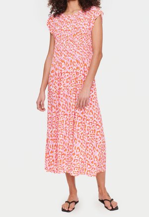 Kvinde iført en midi-kjole med leopardprint i pink og orange med kappelærmer og sorte flip-flops, stående mod en ensfarvet baggrund.
