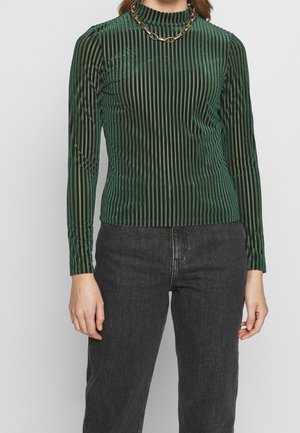 Kobieta ubrana w zielony pasiasty, aksamitny top z długim rękawem, złoty łańcuszek i ciemnoszare dżinsy z wysokim stanem na jednolitym tle.