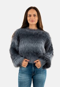 Suéter gris y esponjoso con mangas largas, cuello redondo y una textura suave, combinado con pantalones de mezclilla azules.