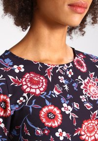 Šaty s květinovým potiskem a kulatým výstřihem. Tkanina je tmavě námořnická modř s červenými, bílými a modrými květy a složitými listovými vzory.