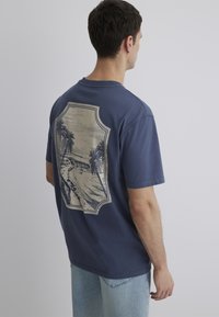 Navy blue short-sleeve t-shirt featuring a light-colored graphic print with palm trees and a beach scene on the back. Cotton material.