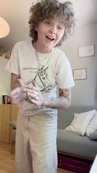 T-shirt branca com uma ilustração de cavalo, colares em camadas, calças largas de cor clara e tatuagens visíveis nos braços.